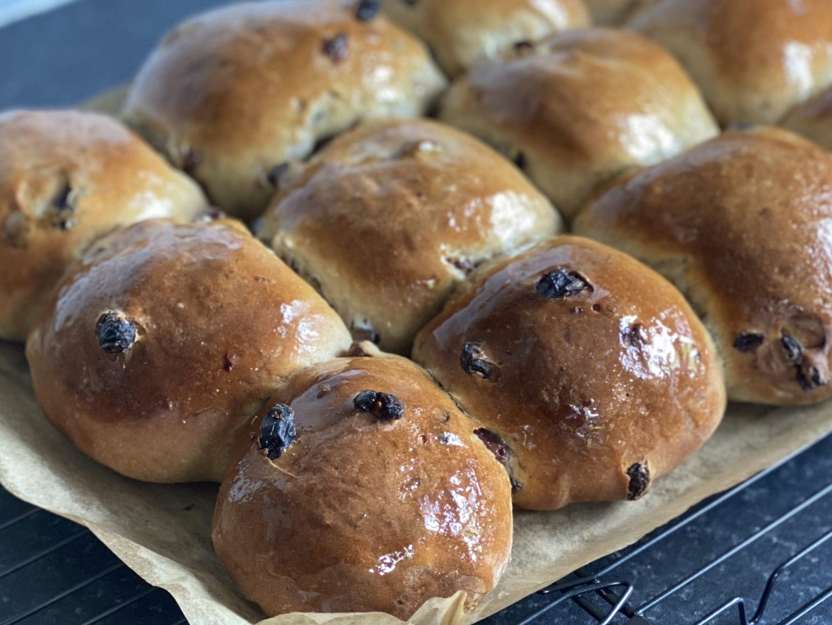 A dozen spiced fruited buns on a cooling rack